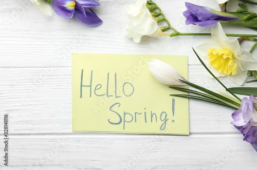 Card with words HELLO SPRING and fresh flowers on white wooden table, flat lay