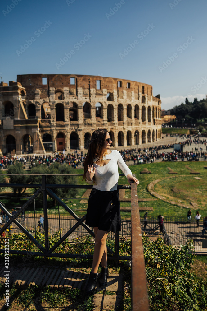 Fashion outdoor photo of beautiful girl with long hair posing near Colosseum in Rome. Young woman looks at Colosseum and people, Rome, Italy. It is top tourist attraction of Rome.