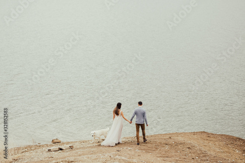 Enamored  wedding couple walk near the lake with a white dog. Rear view of a beautiful wedding couple.