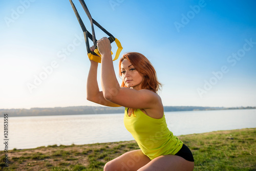 Young woman exercising with suspension trainer sling in park, near the lake.