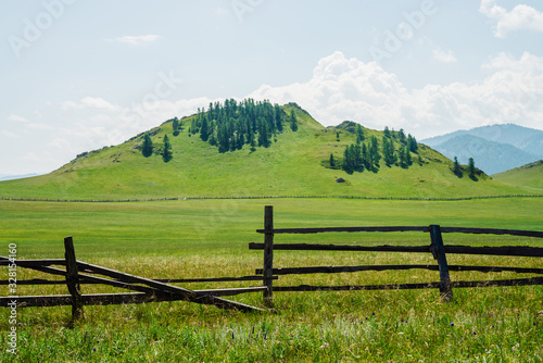 Beautiful sunny landscape with green forest mountain and vast field with long fence. Hill with coniferous trees on top. Vivid scenery with flat field behind wood fence in mountains. Scenic alpine view