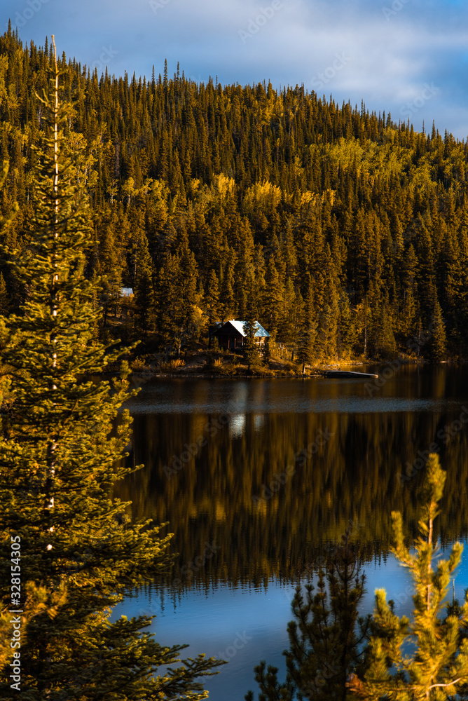 Fototapeta premium Tranquil Secluded Log Cabin on Blue Lake with Reflection In Water Surrounded By Colorful Trees in Alaska Nature Scene