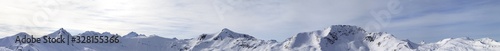 Large panorama of high winter mountains with snowy slopes and sunlit cloudy sky
