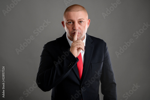 Businessman dressed in black suit placing fingers on lips as if to say shhh.