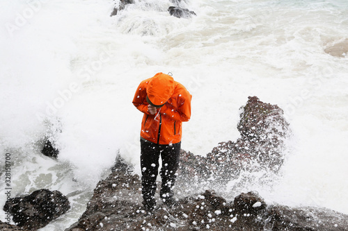 man in waterproof coat standing on rocks at beach splashed by waves
