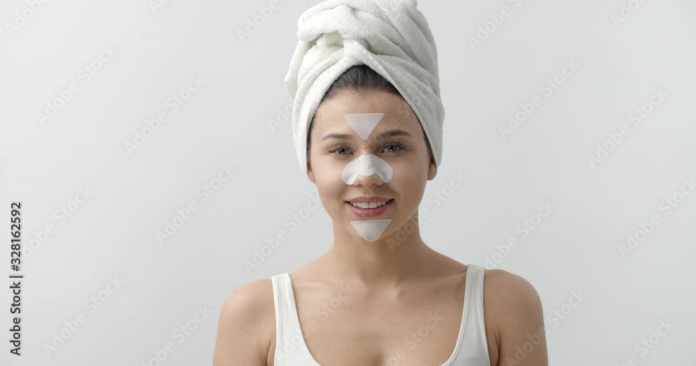 Portrait of happy young girl with bath towel on head looking and smiling on camera with cleansing patches on t-zone. Beautiful woman using cosmetic mask to remove blackheads. Portrait of happy young girl with bath towel on head looking and smiling on camera with cleansing patches on t-zone. Beautiful woman using cosmetic mask to remove blackheads.