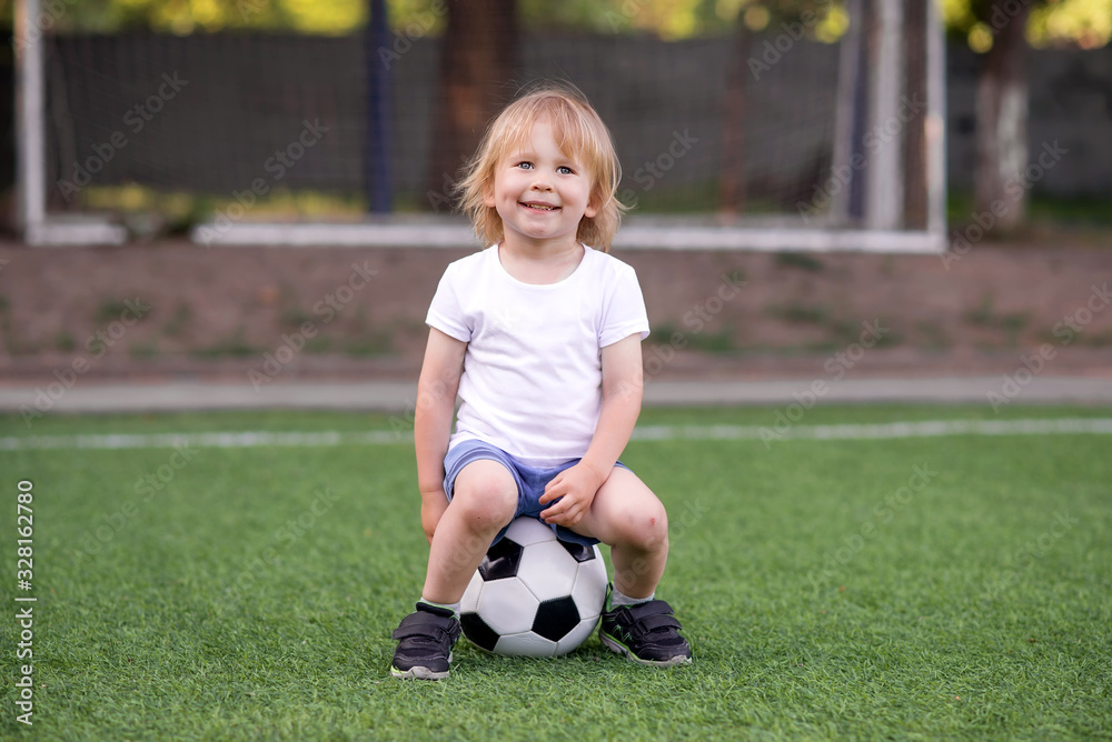 Portrait of blonde toddler kid sitting on soccer ball at football field with broad happy smile. Nets (goalposts) behind. Little sportsman at playground or soccer lesson in kindergarten or preschool. Portrait of blonde toddler kid sitting on soccer ball at football field with broad happy smile. Nets (goalposts) behind. Little sportsman at playground or soccer lesson in kindergarten or preschool.