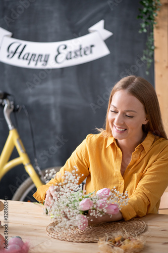 Positive young woman in yellow blouse sitting at table in Easter workshop and making beautiful bouquet from tulips