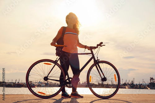 After cycling young red-haired woman stands with bicycle on pier of city seaport against background of ships and cranes at sunset in orange sun, looks into horizon. Lifestyle