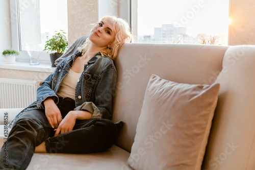 happy lovely blonde girl resting on a sofa in a bright sunny room on the background of a large window © Sergey