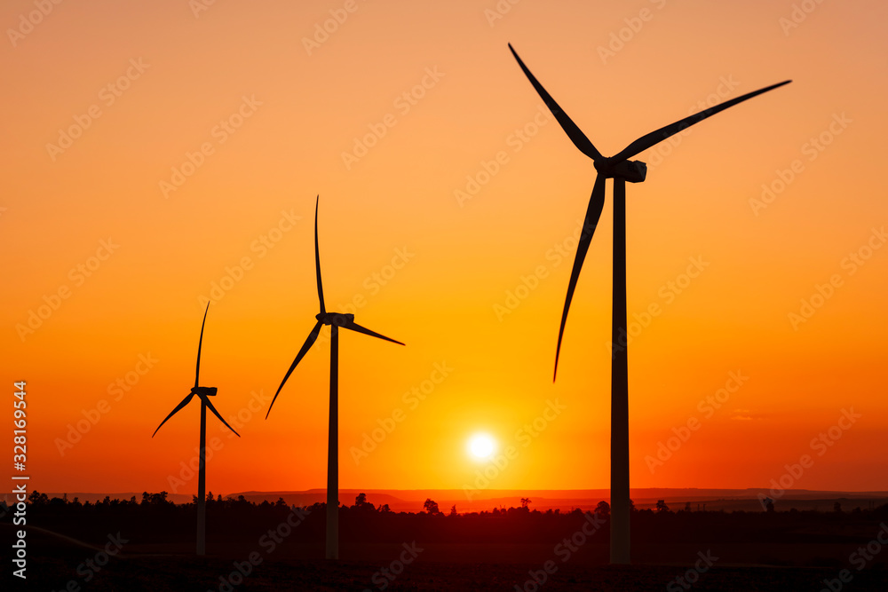 Large wind turbines with blades in field on sky background with bright orange sunset. Silhouettes of windmills, big orange sun disk in summer. Alternative energy sources. Ecological energy