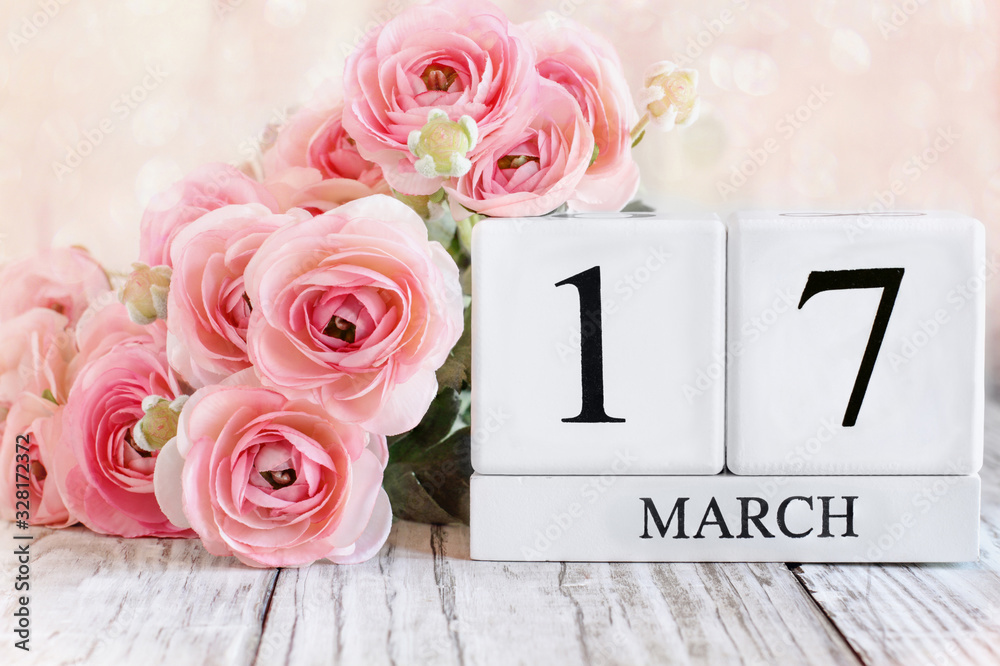Fototapeta premium White wood calendar blocks with the date March 17th and pink ranunculus flowers over a wooden table. Selective focus with blurred background. St Patrick Day.