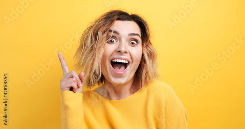 Portrait happy young woman of finding solution Idea pointing forward an index finger on isolated yellow background, looking directly at the camera smiling. Monotone. Emotions of people
