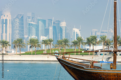 Retro historical boat with blurred panoramic view of modern skyline of Doha and green palms on background