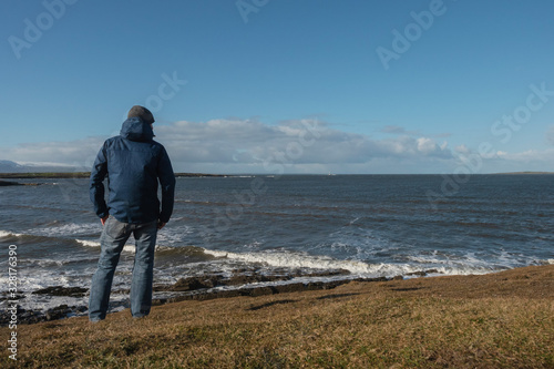 Male tourist by the ocean in warm windproof clothes enjoying landscape, bright sunny day, Rose point, county Sligo, Ireland.