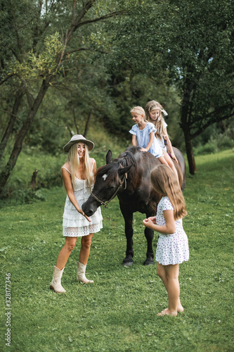 Beautiful young mother in white dress and stylish hat walking with three girls daughters and brown horse outdoors. Two girls riding horse, one sister is feeding the horse with mom. Family with horse