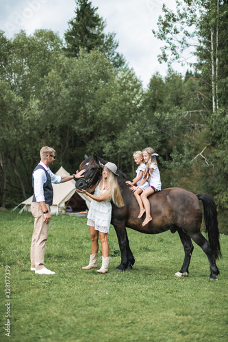 Happy young family with two children, wearing stylish casual boho clothes and beautiful horse in the forest or meadow. Summer camping tent, indian wigwam hut, Tipi on the background