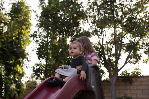 Two girls are helping their toddler baby sister go down a slide in their backyard during summertime in California.