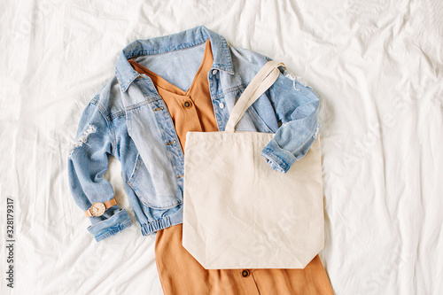 Blue jean jacket and beige dress with tote bag on white bed. Women's stylish autumn outfit. Trendy clothes with white eco bag mockup. Flat lay, top view.