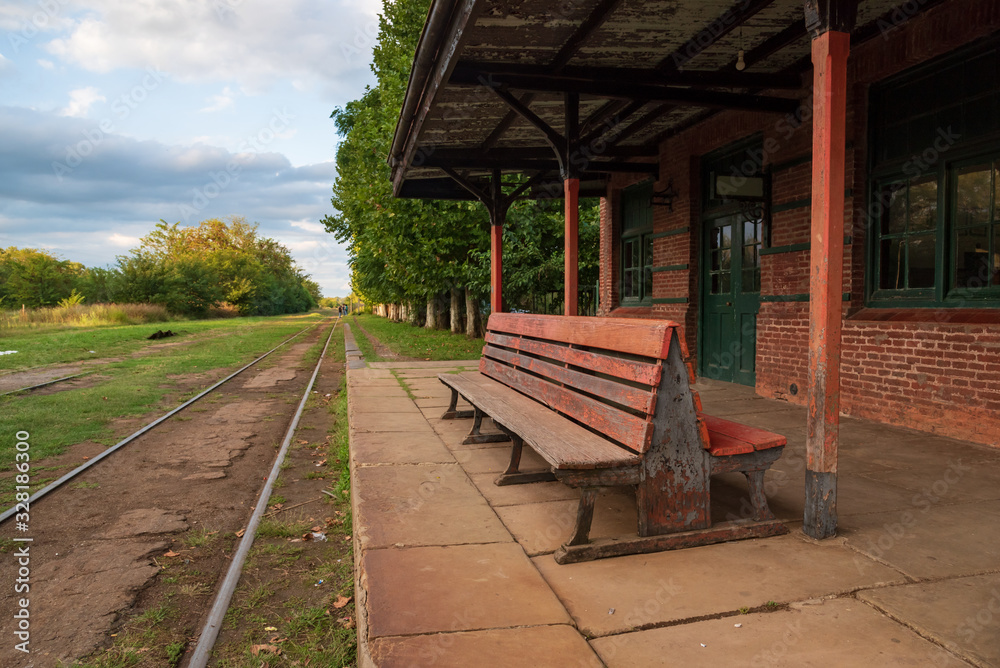 A wooden bench in an old abandoned train station with a view of the railroad with grass aroun it