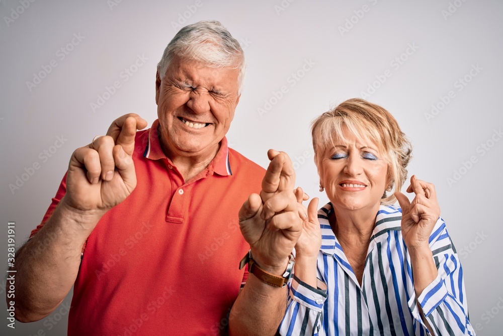 Senior beautiful couple standing together over isolated white background gesturing finger crossed smiling with hope and eyes closed. Luck and superstitious concept. Senior beautiful couple standing together over isolated white background gesturing finger crossed smiling with hope and eyes closed. Luck and superstitious concept.