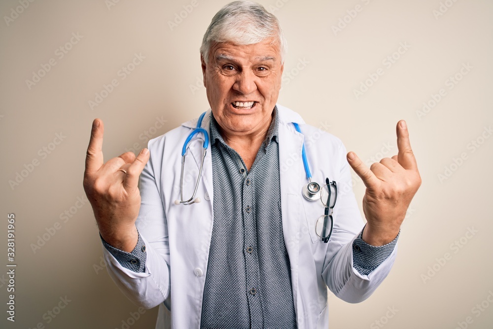 Senior handsome hoary doctor man wearing coat and stethoscope over white background shouting with crazy expression doing rock symbol with hands up. Music star. Heavy concept. Senior handsome hoary doctor man wearing coat and stethoscope over white background shouting with crazy expression doing rock symbol with hands up. Music star. Heavy concept.