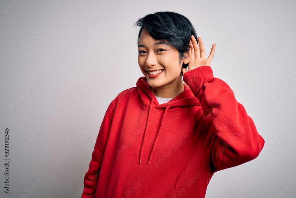 Young beautiful asian girl wearing casual sweatshirt with hoodie over white background smiling with hand over ear listening an hearing to rumor or gossip. Deafness concept. Young beautiful asian girl wearing casual sweatshirt with hoodie over white background smiling with hand over ear listening an hearing to rumor or gossip. Deafness concept.