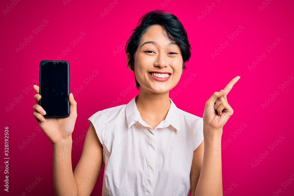 Young beautiful asian girl holding smartphone showing screen over isolated pink background surprised with an idea or question pointing finger with happy face, number one