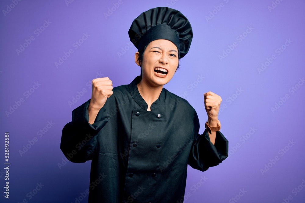 Young beautiful chinese chef woman wearing cooker uniform and hat over purple background angry and mad raising fists frustrated and furious while shouting with anger. Rage and aggressive concept. Young beautiful chinese chef woman wearing cooker uniform and hat over purple background angry and mad raising fists frustrated and furious while shouting with anger. Rage and aggressive concept.