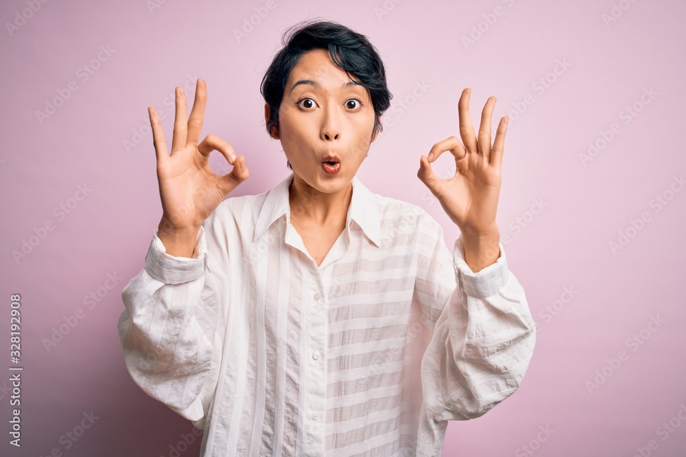 Young beautiful asian girl wearing casual shirt standing over isolated pink background looking surprised and shocked doing ok approval symbol with fingers. Crazy expression Young beautiful asian girl wearing casual shirt standing over isolated pink background looking surprised and shocked doing ok approval symbol with fingers. Crazy expression