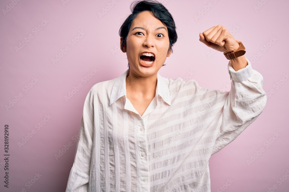 Young beautiful asian girl wearing casual shirt standing over isolated pink background angry and mad raising fist frustrated and furious while shouting with anger. Rage and aggressive concept. Young beautiful asian girl wearing casual shirt standing over isolated pink background angry and mad raising fist frustrated and furious while shouting with anger. Rage and aggressive concept.