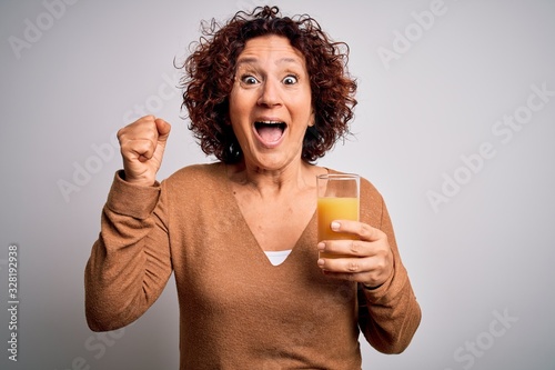 Middle age curly hair woman drinking healthy glass of orange juice over white background screaming proud and celebrating victory and success very excited, cheering emotion © Krakenimages.com