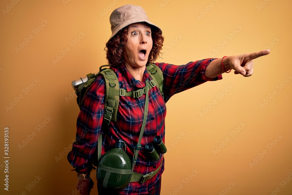 Middle age curly hair hiker woman hiking wearing backpack and water canteen using binoculars Pointing with finger surprised ahead, open mouth amazed expression, something on the front
