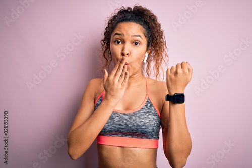 Beautiful sportswoman with curly hair doing sport wearing sportswear using smartwatch cover mouth with hand shocked with shame for mistake, expression of fear, scared in silence, secret concept © Krakenimages.com
