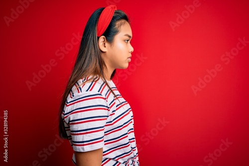 Young beautiful asian girl wearing casual striped t-shirt over isolated red background looking to side, relax profile pose with natural face and confident smile. © Krakenimages.com