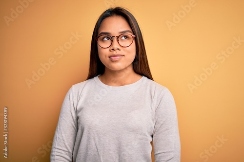 Young beautiful asian girl wearing casual sweater and glasses over yellow background smiling looking to the side and staring away thinking.
