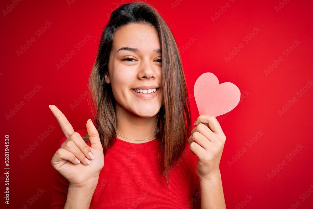 Young beautiful brunette romantic girl holding red paper heart shape over isolated background very happy pointing with hand and finger to the side Young beautiful brunette romantic girl holding red paper heart shape over isolated background very happy pointing with hand and finger to the side