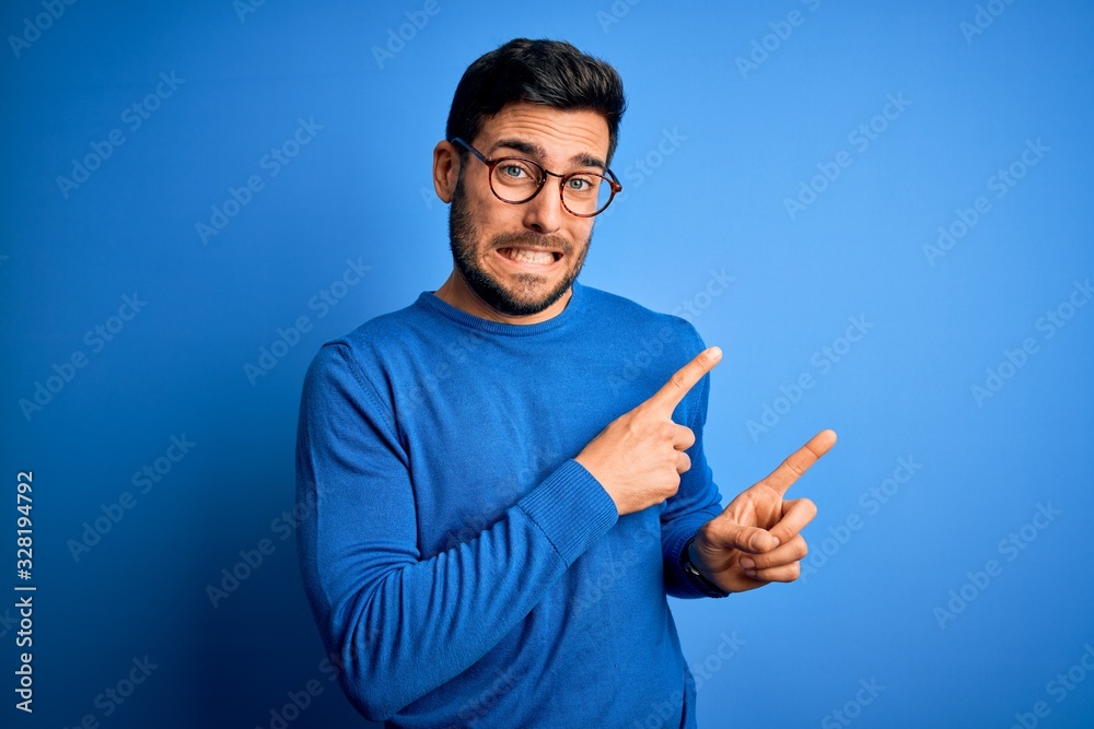 Young handsome man with beard wearing casual sweater and glasses over blue background Pointing aside worried and nervous with both hands, concerned and surprised expression Young handsome man with beard wearing casual sweater and glasses over blue background Pointing aside worried and nervous with both hands, concerned and surprised expression