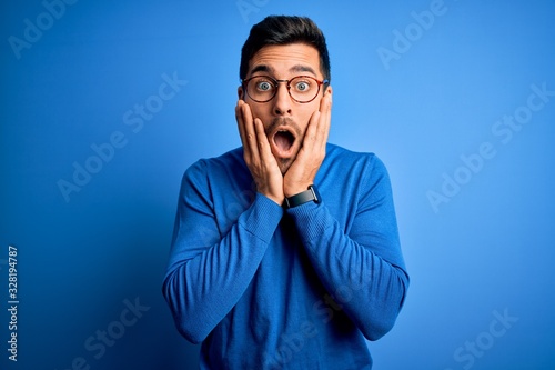 Young handsome man with beard wearing casual sweater and glasses over blue background afraid and shocked, surprise and amazed expression with hands on face photo