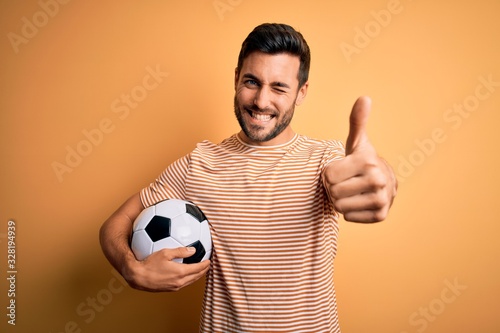 Handsome player man with beard playing soccer holding footballl ball over yellow background approving doing positive gesture with hand, thumbs up smiling and happy for success. Winner gesture.