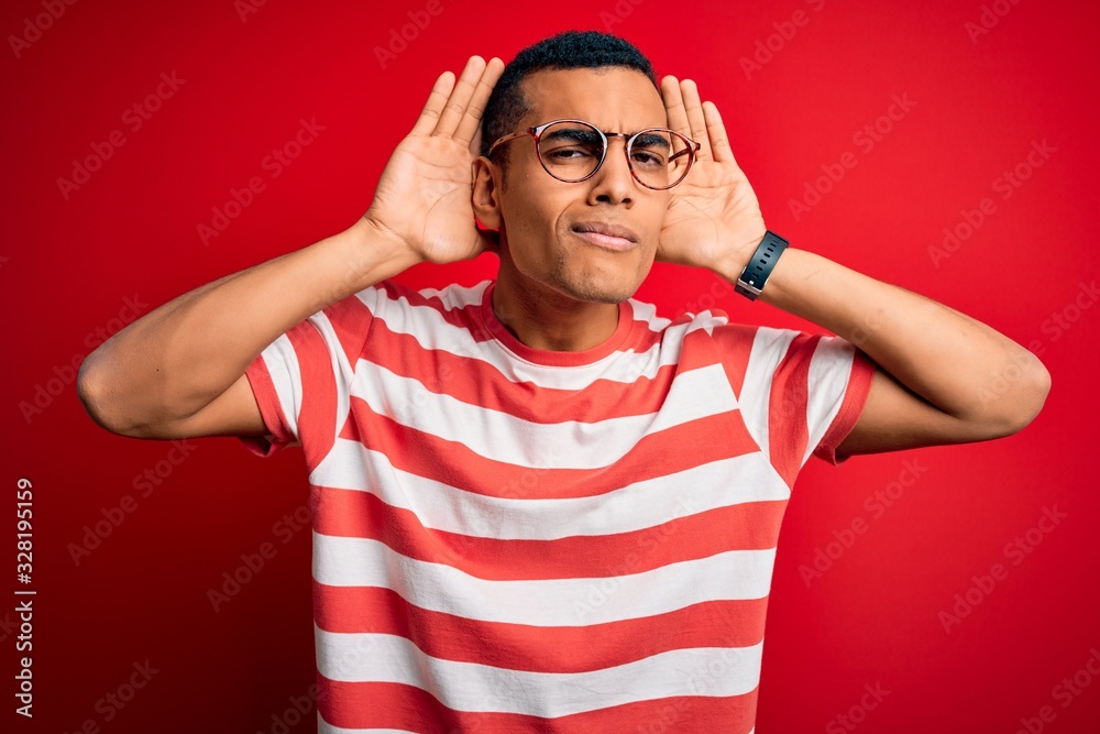 Young handsome african american man wearing casual striped t-shirt and glasses Trying to hear both hands on ear gesture, curious for gossip. Hearing problem, deaf Young handsome african american man wearing casual striped t-shirt and glasses Trying to hear both hands on ear gesture, curious for gossip. Hearing problem, deaf