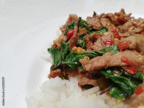 Popular spicy Thai dish, jasmin rice topped with stir fried beef with basil leaves, chilli and garic on white plate.