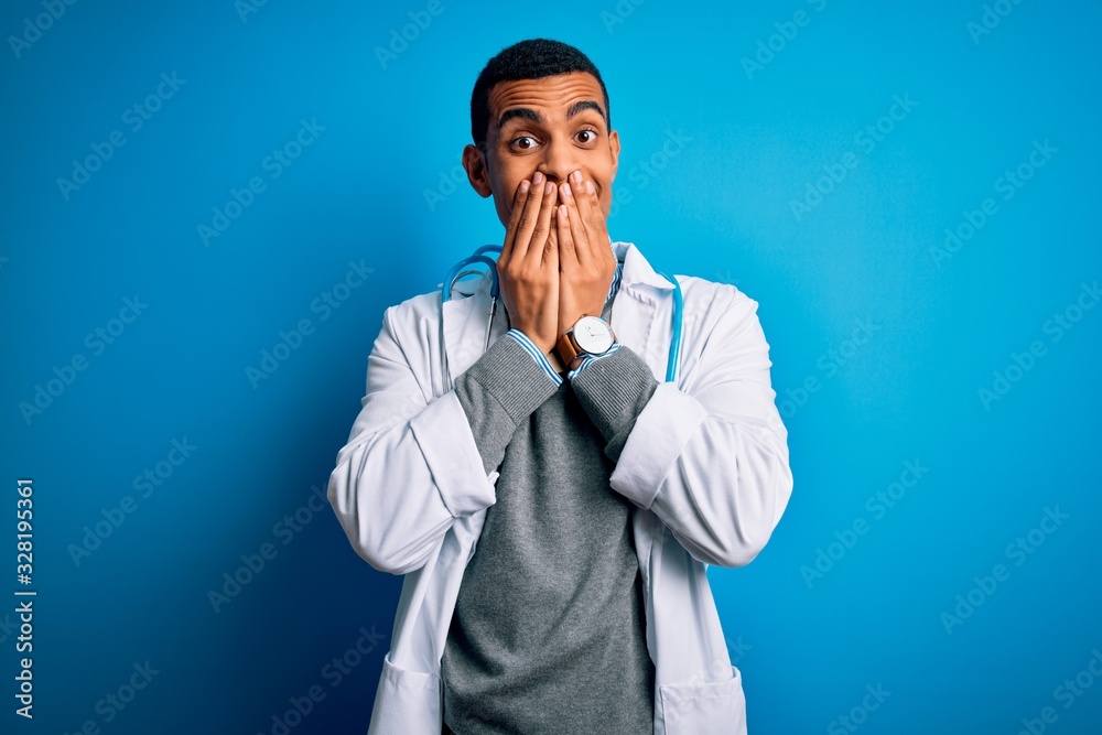 Handsome african american doctor man wearing coat and stethoscope over blue background laughing and embarrassed giggle covering mouth with hands, gossip and scandal concept