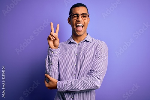 Handsome african american man wearing striped shirt and glasses over purple background smiling with happy face winking at the camera doing victory sign with fingers. Number two.