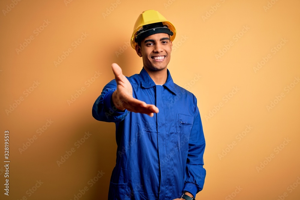 Young handsome african american worker man wearing blue uniform and security helmet smiling cheerful offering palm hand giving assistance and acceptance.