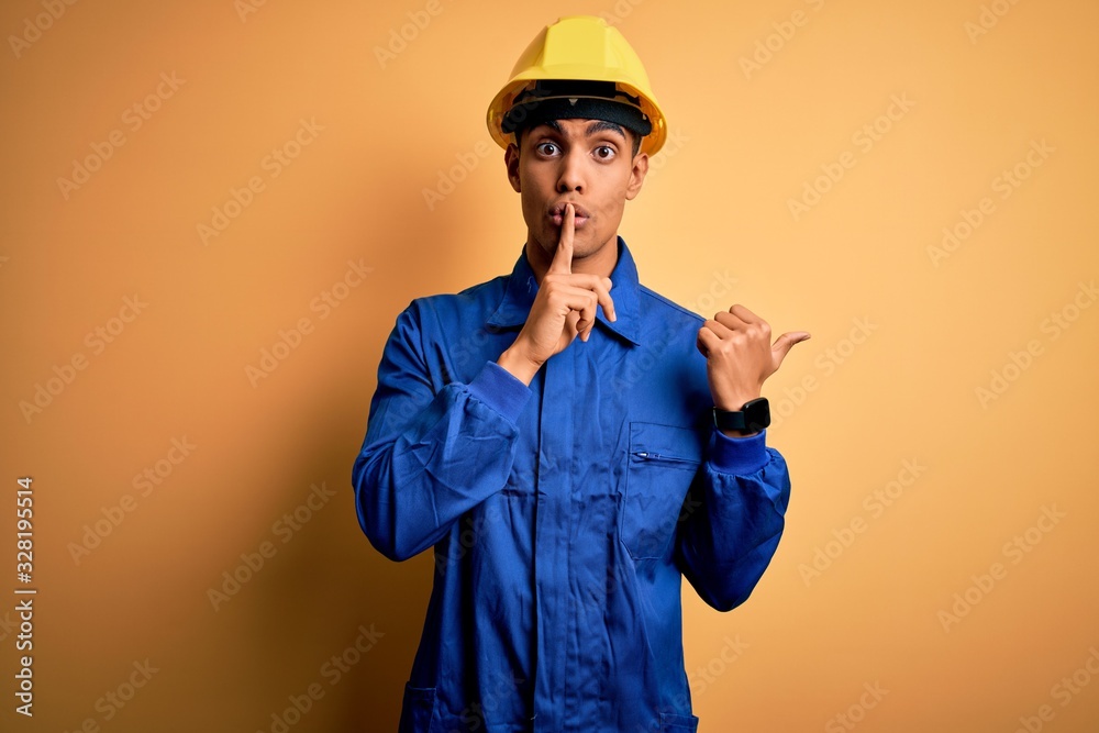 Young handsome african american worker man wearing blue uniform and security helmet asking to be quiet with finger on lips pointing with hand to the side. Silence and secret concept.