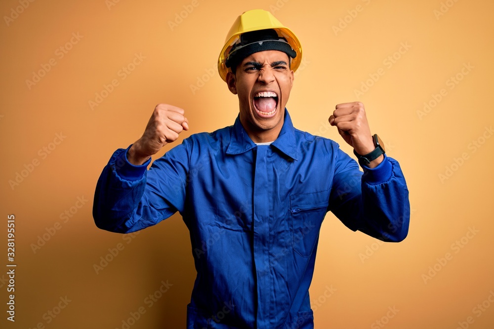 Young handsome african american worker man wearing blue uniform and security helmet angry and mad raising fists frustrated and furious while shouting with anger. Rage and aggressive concept.
