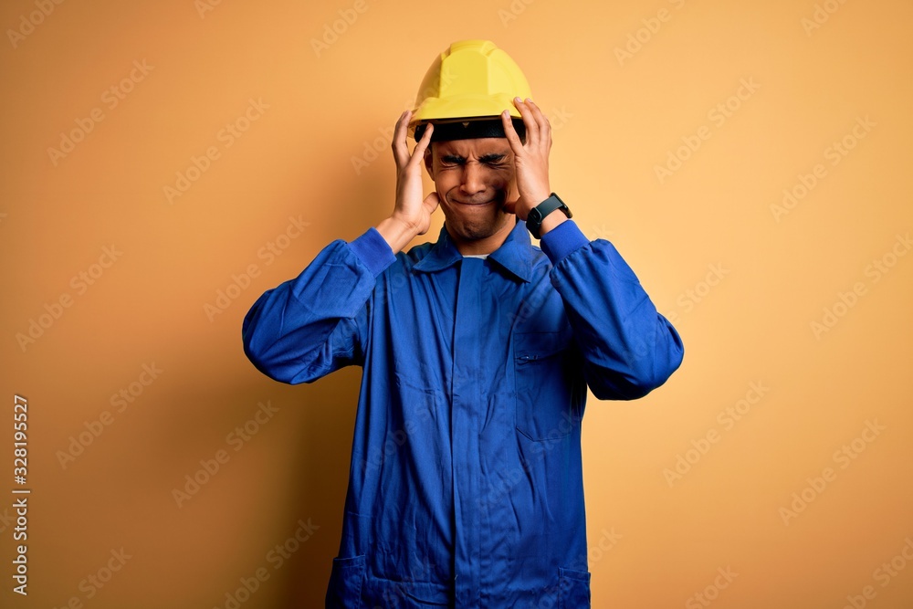 Young handsome african american worker man wearing blue uniform and security helmet suffering from headache desperate and stressed because pain and migraine. Hands on head.