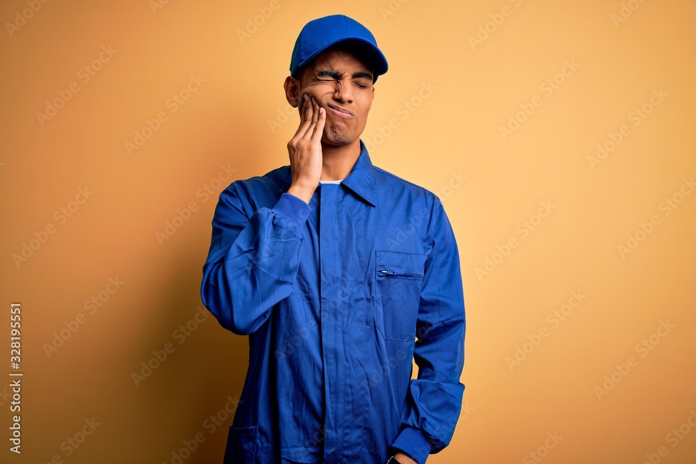 Young african american mechanic man wearing blue uniform and cap over yellow background touching mouth with hand with painful expression because of toothache or dental illness on teeth. Dentist