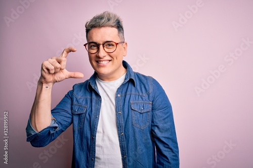 Young handsome modern man wearing glasses and denim jacket over pink isolated background smiling and confident gesturing with hand doing small size sign with fingers looking and the camera. Measure